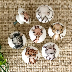May include: Set of seven ceramic knobs featuring various animal designs, including a zebra, giraffe, lion, elephant, hippopotamus, and leopard. The knobs are white with colorful animal illustrations.