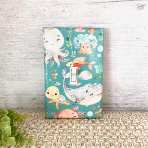 May include: A light switch cover with a teal background and a whimsical underwater scene featuring an octopus, a whale, a turtle, a fish, and coral.