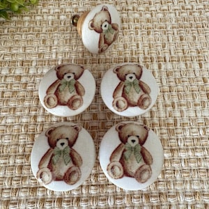 May include: Five round cabinet knobs featuring a printed illustration of a brown teddy bear with a light green bow tie. Each knob has a cream-colored fabric-like surface and a metal screw base, showcasing a consistent design across all pieces.