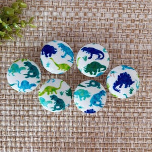 May include: Six white fabric buttons with a blue and green dinosaur pattern. The buttons are round and have a white background with blue and green dinosaur silhouettes.