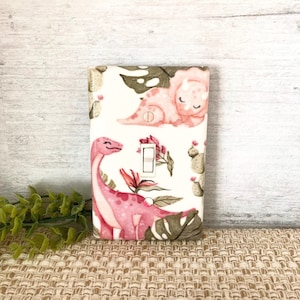 May include: A light switch cover with a pink dinosaur and cactus pattern. The cover is white with a pink dinosaur and cactus pattern. The switch is white.