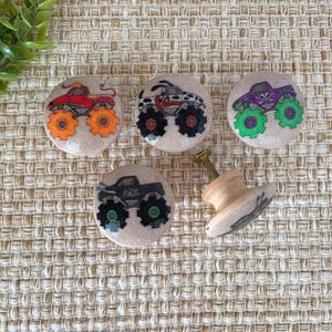 May include: Set of five decorative cabinet knobs featuring monster truck designs. The knobs have a light beige background with colorful monster truck illustrations in red, black, purple, and gray. The knobs are round with a wooden base and a screw for installation.