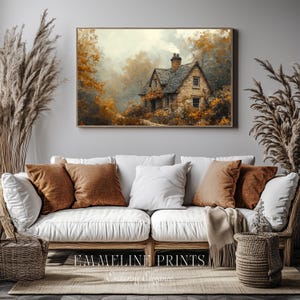 May include: A framed print of a stone cottage nestled in a forest with autumn foliage. The artwork is displayed above a white sofa with brown and white pillows. The text "EMMELINE PRINTS" and "Enduring Elegance" is at the bottom.