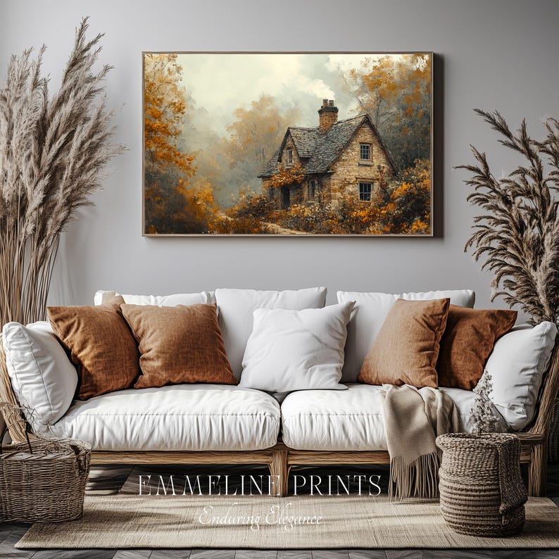 May include: A framed print of a stone cottage nestled in a forest with autumn foliage. The artwork is displayed above a white sofa with brown and white pillows. The text "EMMELINE PRINTS" and "Enduring Elegance" is at the bottom.
