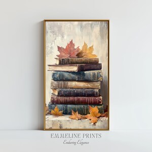 Puede incluir: Impresión enmarcada que muestra una pila de libros antiguos con cubiertas desgastadas en tonos marrón, azul y burdeos. Hojas de otoño rojas y doradas descansan sobre y alrededor de los libros. El texto "EMMELINE PRINTS Enduring Elegance" está en la parte inferior.