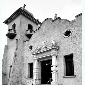 Może przedstawiać: Czarno-białe zdjęcie budynku kościoła z wysoką wieżą i dużym wejściem. Budynek ma białe tynki zewnętrzne i krzyż na szczycie wieży.