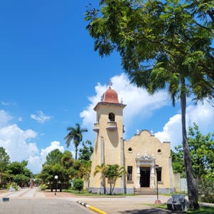 Może przedstawiać: Żółty kościół z czerwonym dachem i wieżą z dzwonem. Kościół otoczony jest drzewami i błękitnym niebem z białymi chmurami.