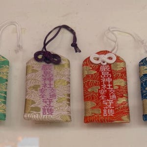 May include: Four Japanese omamori charms in green, gold, red, and blue. Each charm has a loop for hanging and features Japanese text. The charms have a decorative wave pattern.