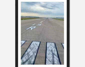 May include: A framed photograph of an airport runway featuring the white painted text 15 R on the gray asphalt surface. The runway includes white rectangular markings with visible tire skid marks, leading toward a distant horizon under a cloudy sky.