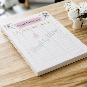 Könnte beinhalten: Ein weißes Parent Contact Log Notizbuch mit einem rosa Banner und floralen Akzenten. Das Notizbuch hat ein Rasterlayout zur Aufzeichnung von Informationen. Der Text "Made by Misa" ist kursiv geschrieben. Im Hintergrund steht eine kleine Vase mit weißen Blumen.