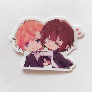 May include: A sticker featuring two anime characters, one with pink hair and the other with brown hair, both wearing suits. The character with pink hair is holding the other character's arm. There are two pink hearts in the top right corner of the sticker.