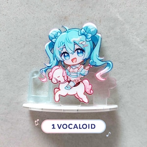 Op de afbeelding: Een acryl display met een chibi-stijl personage met lang blauwgroen haar in staartjes, gekleed in een lichtblauwe en roze outfit, rijdend op een wit paard. Het personage heeft grote blauwe ogen en een glimlach. De tekst 1 VOCALOID is zichtbaar.