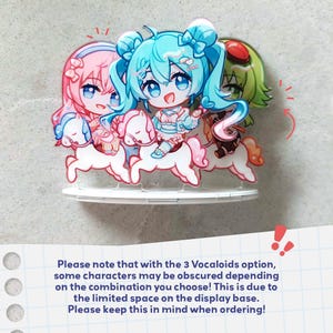 Op de afbeelding: Een acryl displaystandaard met drie chibi-stijl figuren met roze, groenblauw en groen haar, elk rijdend op een wit carrouselpaard. De figuren zijn geplaatst op een ronde witte voet, inclusief een opmerking over de positionering van de figuren.