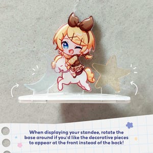 Op de afbeelding: Een acryl display met een blond personage met een bruine strik op een wit paard, geflankeerd door een doorschijnende halve maan en een ster. De voet is wit, met de tekst: When displaying your standee, rotate the base around.