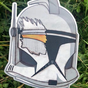May include: A sticker featuring a detailed illustration of a Clone Trooper helmet. The helmet is primarily white and gray, with black accents and a yellow visor. The design includes battle damage and a stylized antenna.