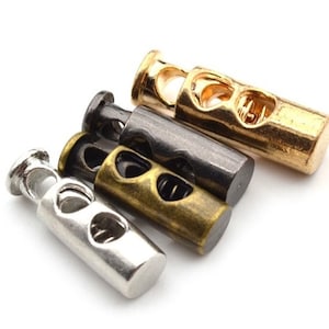 May include: Four metal cord stoppers in different finishes: gold, black, bronze, and silver. Each stopper has a cylindrical shape with two heart-shaped cutouts and a spring mechanism.