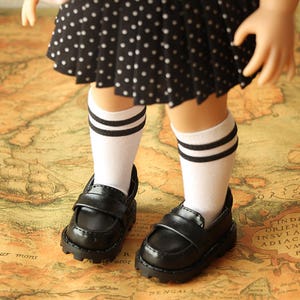 May include: A pair of black leather doll shoes with white socks that have black stripes. The shoes have a buckle closure and a chunky sole. The doll is wearing a black and white polka dot skirt.
