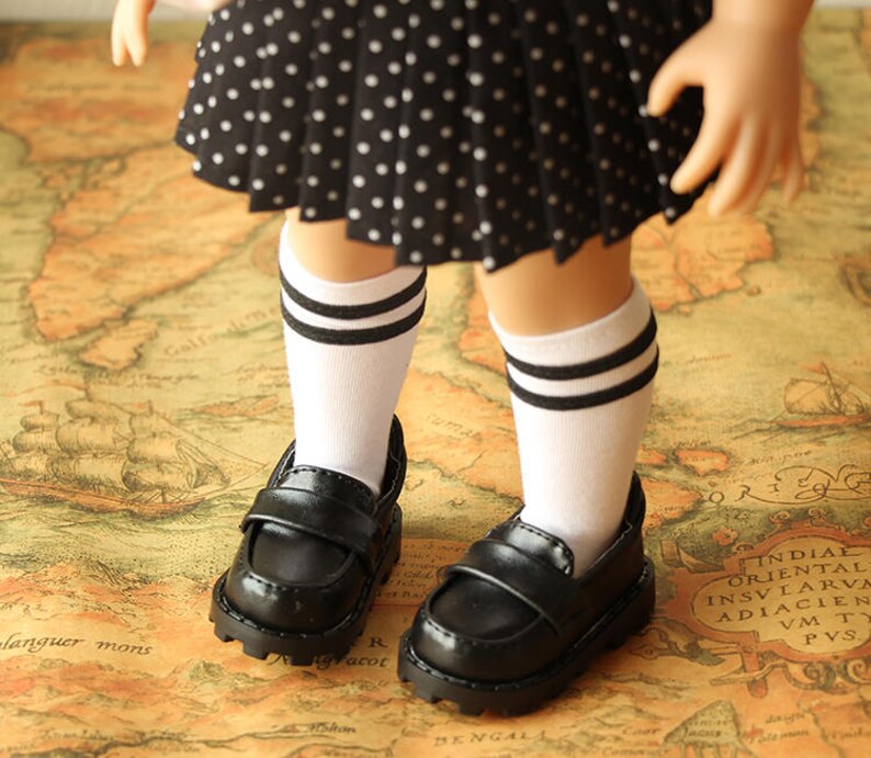 May include: A pair of black leather doll shoes with white socks that have black stripes. The shoes have a buckle closure and a chunky sole. The doll is wearing a black and white polka dot skirt.