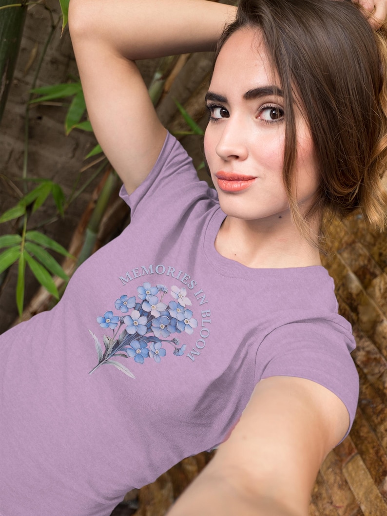May include: A lilac t-shirt with a watercolour illustration of blue forget-me-not flowers. The text "Memories in Bloom" is arched above the floral design. The shirt is made of a soft material.