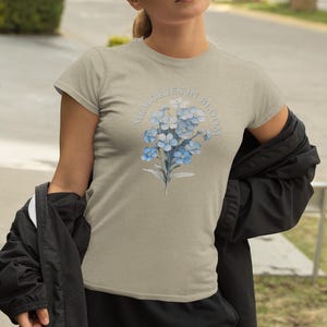 May include: A light tan t-shirt with a floral design of blue flowers and the text "Memories in Bloom". The shirt has short sleeves and a crew neck. The design is centered on the chest.