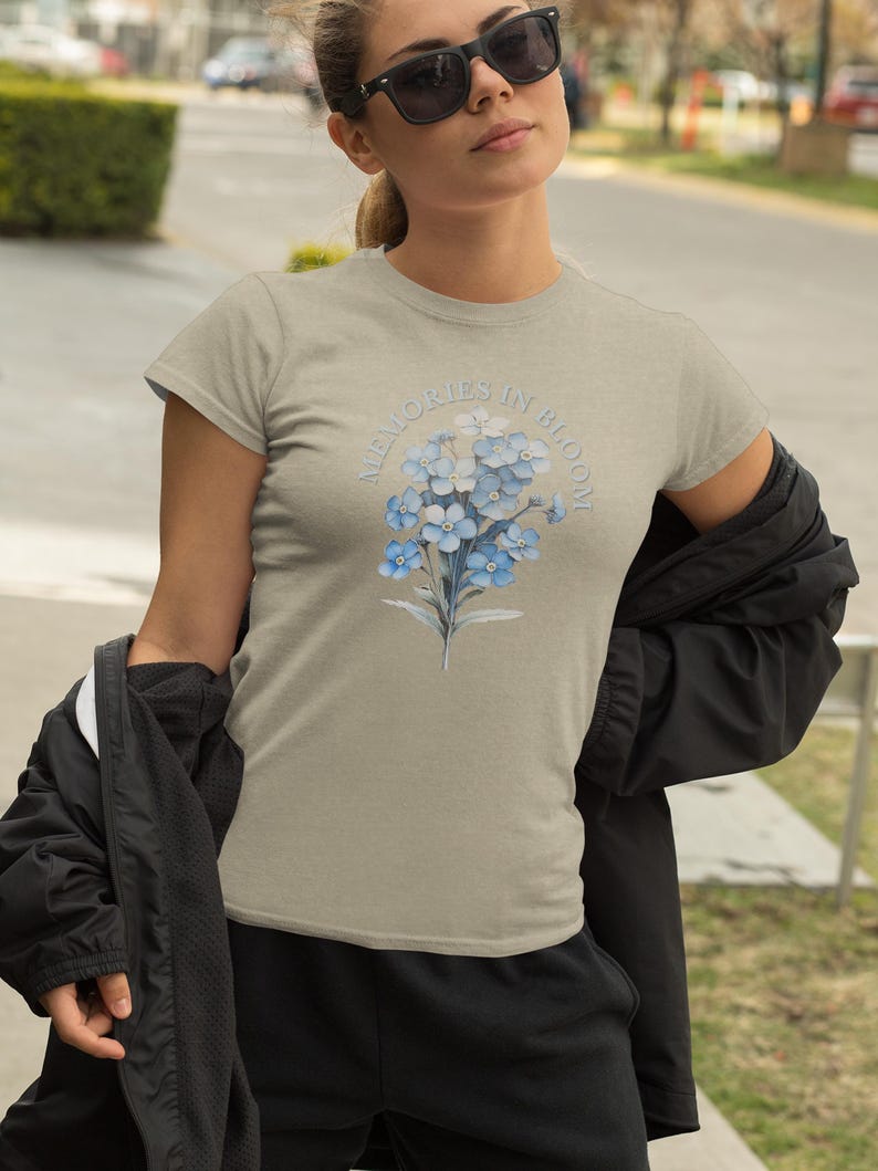 May include: A light tan t-shirt with a floral design of blue flowers and the text "Memories in Bloom". The shirt has short sleeves and a crew neck. The design is centered on the chest.
