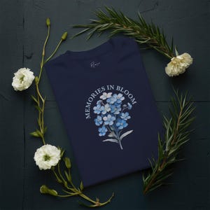 May include: A navy blue t-shirt with a floral design of blue forget-me-nots and the text "MEMORIES IN BLOOM". The shirt is decorated with white flowers and green foliage on a dark background.