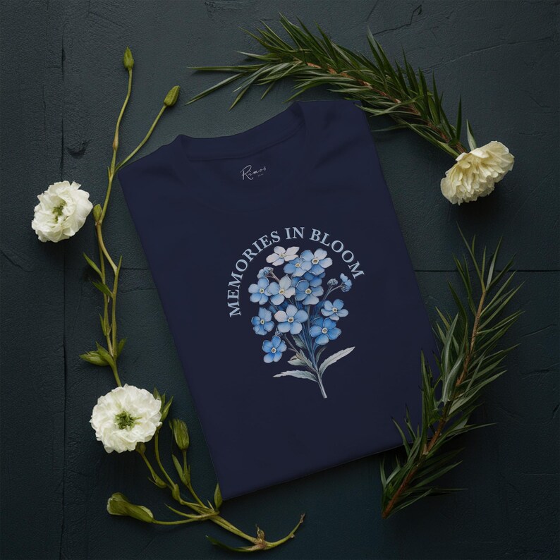 May include: A navy blue t-shirt with a floral design of blue forget-me-nots and the text "MEMORIES IN BLOOM". The shirt is decorated with white flowers and green foliage on a dark background.