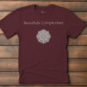 Peut inclure: Un t-shirt bordeaux avec le texte "Beautifully Complicated" au-dessus d'un motif mandala blanc détaillé. Le t-shirt est fait d'une matière douce et est présenté sur une surface en bois.