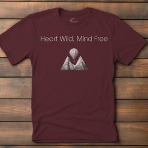 Puede incluir: Camiseta color burdeos con un gráfico blanco de un globo aerostático volando sobre montañas. El texto "Heart Wild, Mind Free" está impreso en la camiseta.