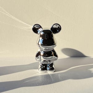 May include: A silver, metallic, cartoon-style figurine of a mouse with large ears. The figurine is standing on a white surface.