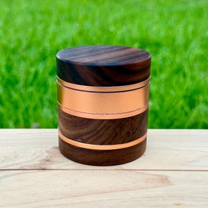 May include: A cylindrical wooden grinder with a dark brown finish and copper-colored bands. The grinder is sitting on a light-colored wooden surface with a green background.