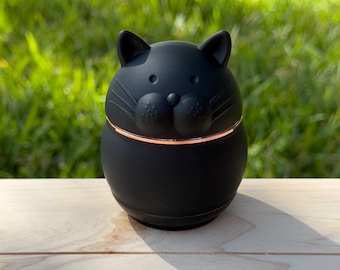 May include: Black ceramic cat-shaped jar with a gold rim. The jar has a cute, cartoon-like face with whiskers.