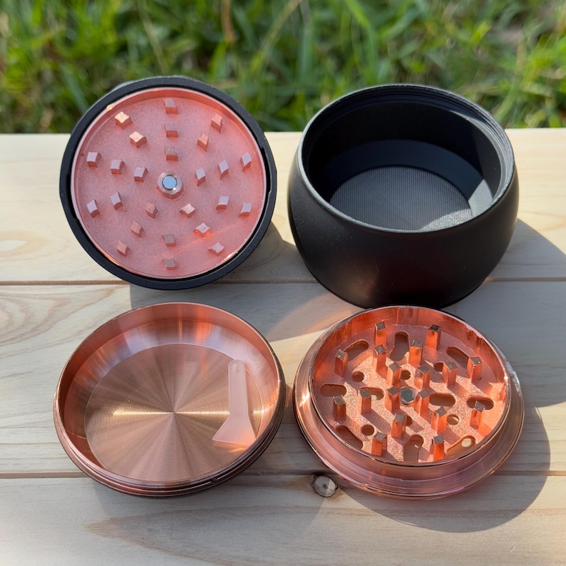 May include: A four-piece grinder with a rose gold finish. The grinder has a black lid, a black base, and two rose gold pieces with teeth for grinding.