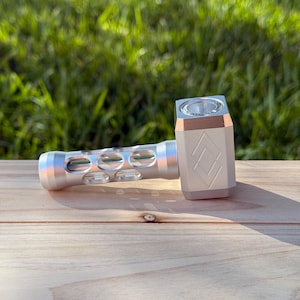 May include: A silver metal pipe shaped like a hammer with a clear glass bowl. The pipe has a diamond-shaped logo on the side.