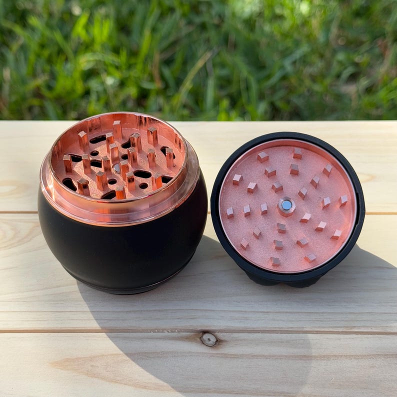 May include: A black and rose gold grinder with a round shape and a magnetic lid. The grinder has a metal top with a series of teeth for grinding herbs.