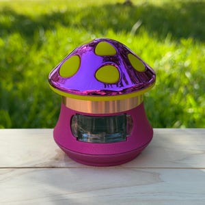 May include: A pink and yellow mushroom-shaped ceramic jar with a clear glass window. The jar has a shiny, metallic finish.