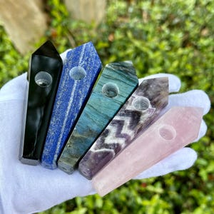 May include: Five gemstone pipes in a variety of colors are displayed. The pipes are black, blue, teal, purple, and pink. Each pipe has a cylindrical hole. The pipes are held in a white gloved hand against a green background.