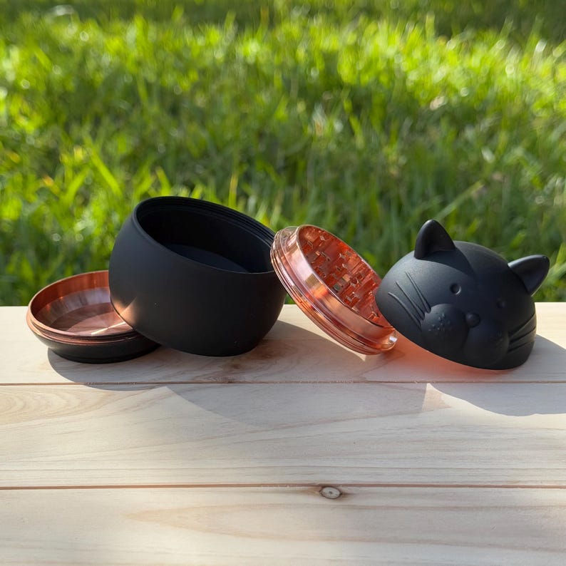 May include: A black and copper grinder with a cat face design. The grinder is in three pieces, with the top piece being the cat face, the middle piece being the grinding mechanism, and the bottom piece being a catch for the ground herbs.