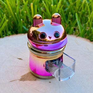 May include: A metallic, iridescent pink and gold herb grinder shaped like a cat head with two small ears on top. The grinder features a clear, rectangular plastic drawer extending from the front, revealing a fine mesh screen inside the cylindrical body.
