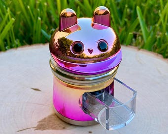 May include: A metallic, iridescent pink and gold herb grinder shaped like a cat head with two small ears on top. The grinder features a clear, rectangular plastic drawer extending from the front, revealing a fine mesh screen inside the cylindrical body.