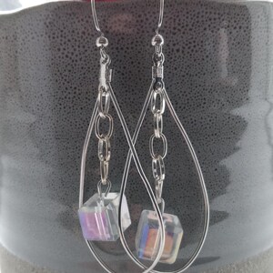Könnte beinhalten: Silberne Tropfenohrringe mit Kettenakzenten und schillernden Würfelanhängern. Die Ohrringe hängen an silbernen Haken und werden in einem grauen Keramikbehälter mit einer roten Blume im Hintergrund präsentiert.