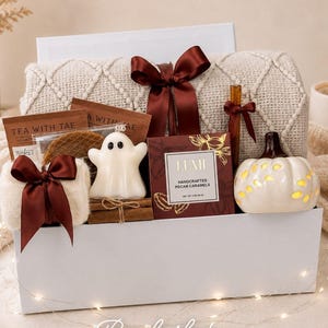 May include: A white gift box containing a cream-colored knit blanket with a dark red ribbon, a ghost-shaped candle, a box of Lumi handcrafted pecan caramels, a tea packet, a small white pumpkin-shaped light, and a cinnamon stick tied with ribbon.
