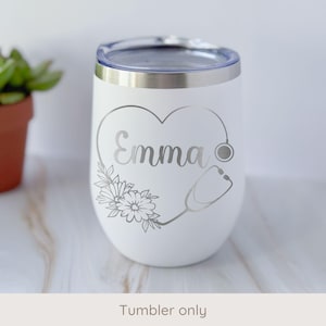 May include: White insulated tumbler with a silver heart-shaped design, the name "Emma", flowers, and a stethoscope. The tumbler has a silver rim and a clear lid. The text "Tumbler only" is at the bottom.