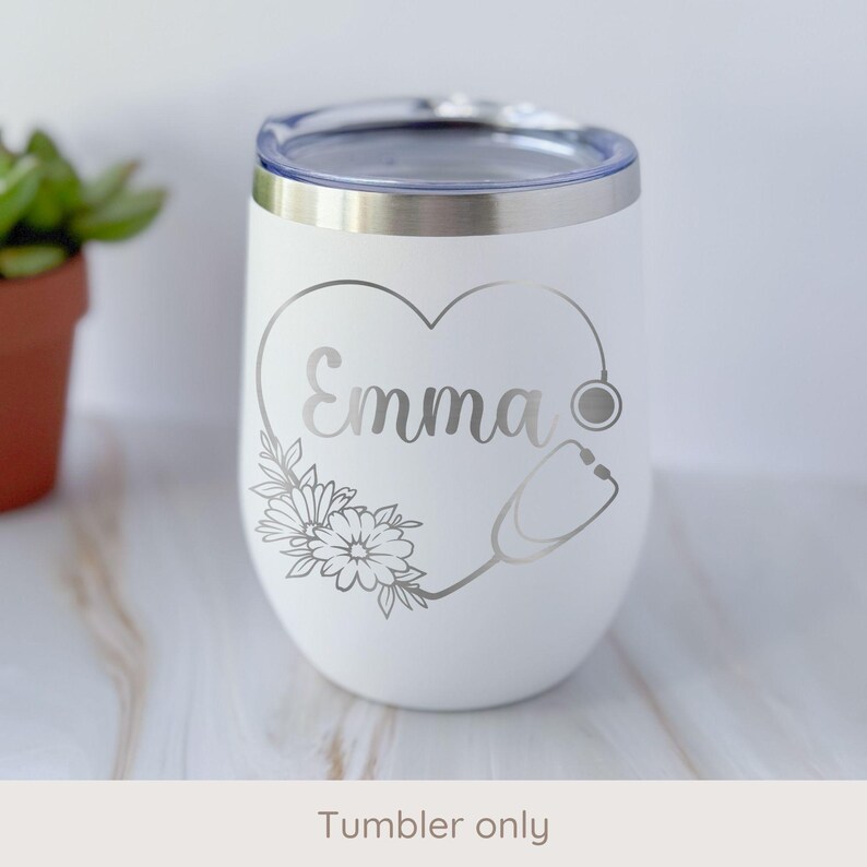 May include: White insulated tumbler with a silver heart-shaped design, the name "Emma", flowers, and a stethoscope. The tumbler has a silver rim and a clear lid. The text "Tumbler only" is at the bottom.