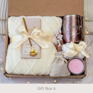 May include: A gift box filled with a cream-colored blanket, chocolate bar, gold spoon, rose gold tumbler with the name "Emma", a small candle, and other treats. The box is tied with a cream ribbon. The text "Gift Box 6" is at the bottom.