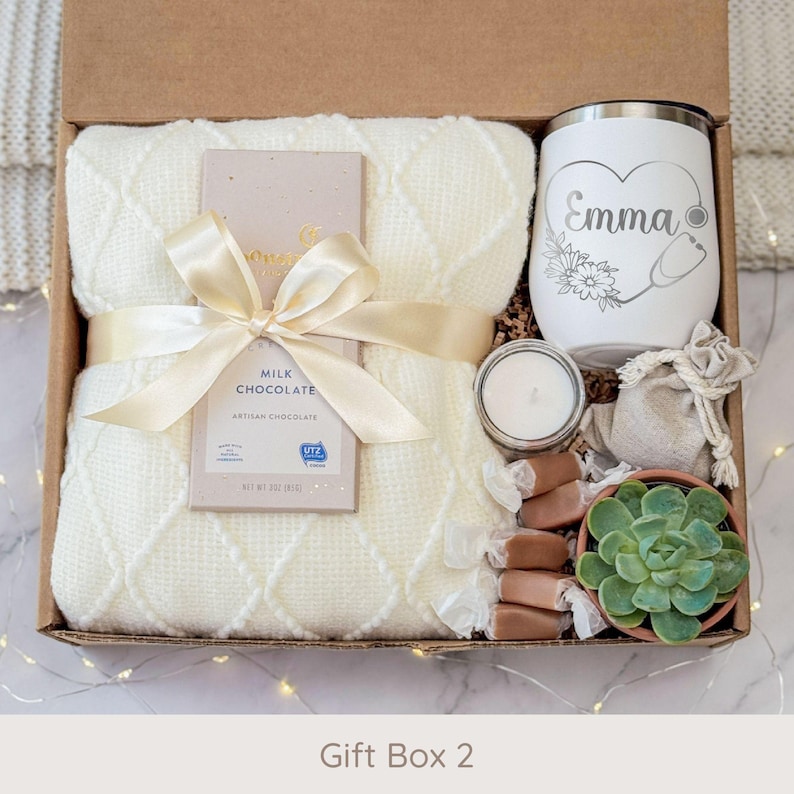 May include: A gift box filled with a cream-colored blanket, milk chocolate bar, white tumbler with a heart and stethoscope design, candle, candies, and a small succulent plant. The box is tied with a cream ribbon.