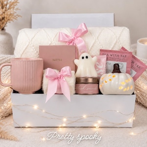 May include: A gift box containing a ribbed pink mug, a dusty rose notebook labeled NOTES, a white ghost-shaped candle, a small pumpkin-shaped ceramic piece, a candle tin labeled blush vanilla, and tea packets, all accented with pink satin ribbons.