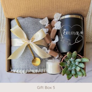 May include: A gift box with a gray blanket tied with a gold ribbon and spoon, a black tumbler with "Emma" and a stethoscope design, wrapped candies, a small candle, and a succulent plant.