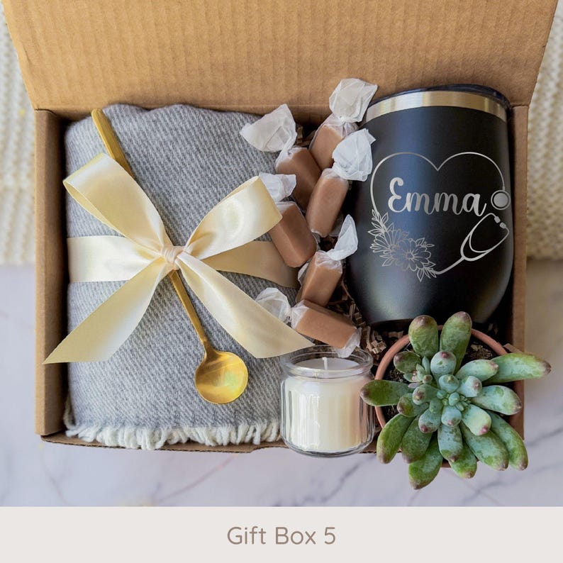 May include: A gift box with a gray blanket tied with a gold ribbon and spoon, a black tumbler with "Emma" and a stethoscope design, wrapped candies, a small candle, and a succulent plant.