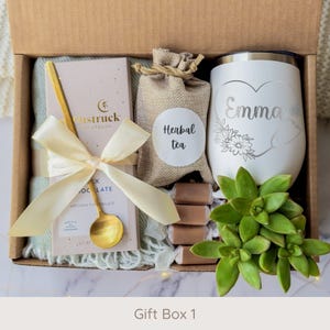 May include: A gift box filled with a variety of items. Includes a white tumbler with the name "Emma" and a floral design, a bar of chocolate, a gold spoon, herbal tea, and a succulent plant. The box is tied with a cream-colored ribbon. The text "Gift Box 1" is at the bottom.